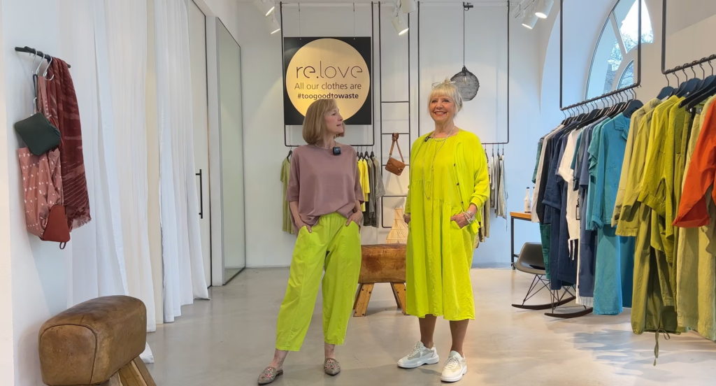 Two women pose in a brightly lit boutique, one wearing a pink top with lime trousers, the other in a bright yellow midi dress.