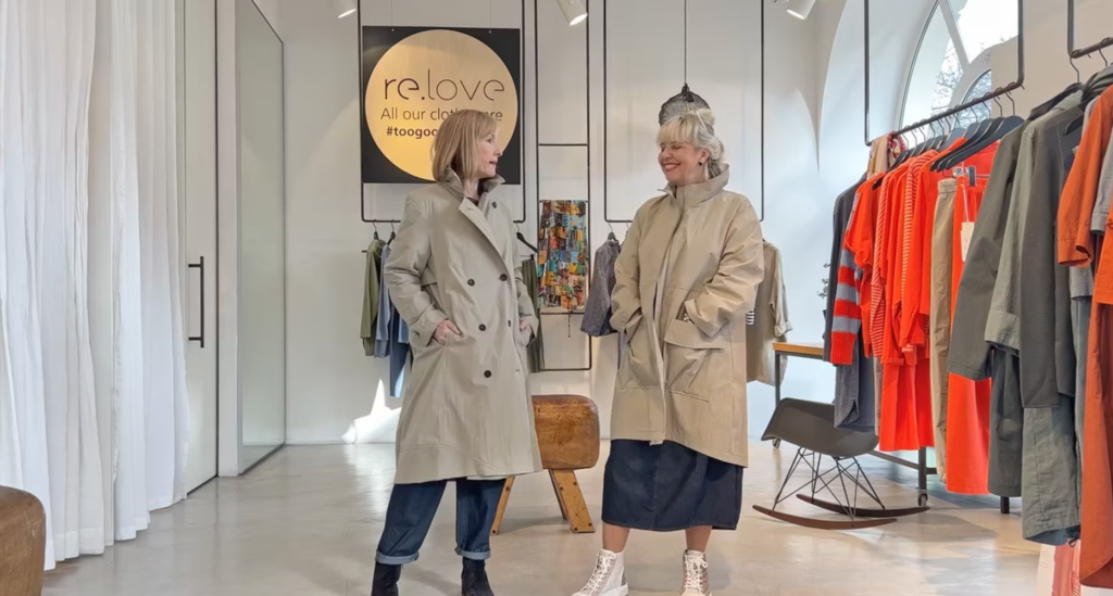 Two women wearing beige coats in a clothing store with racks of brightly colored garments.