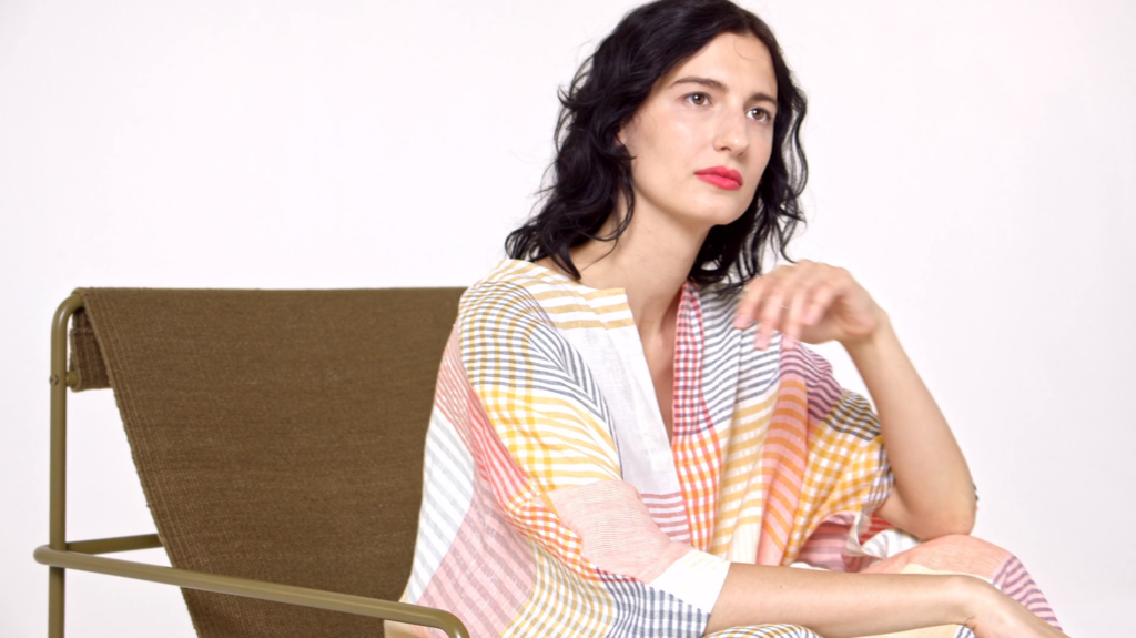 A dark-haired woman sits in an olive green chair wearing a colourful checkered tunic.