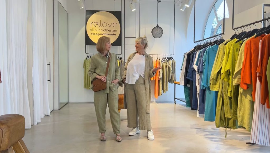 Two women in sage green linen sets stand in a clothing boutique with racks of colorful garments.