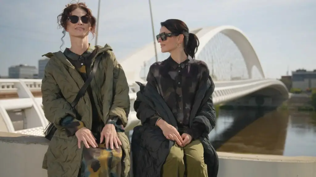 Deux femmes portant des lunettes de soleil et des manteaux sont assises à l'extérieur près d'un pont moderne.