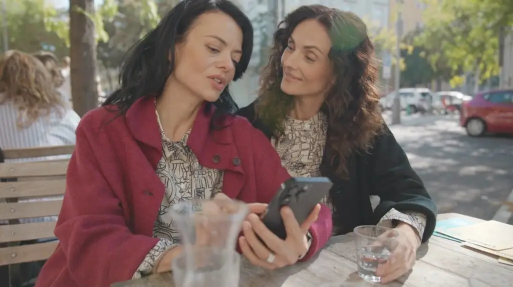 Deux femmes portant des hauts à motifs et des manteaux sombres sont assises ensemble à une table de café en plein air, en train de regarder un téléphone.