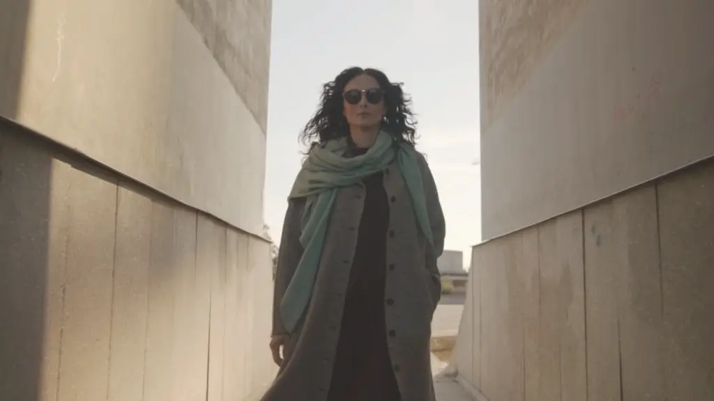 Une femme portant un manteau gris et une écharpe marche entre des murs en pierre.