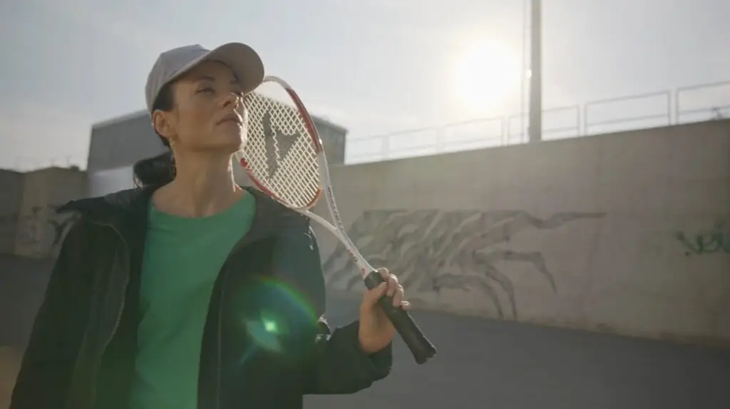 Une femme portant une casquette grise, un haut vert et une veste foncée tient une raquette de tennis à l'extérieur.