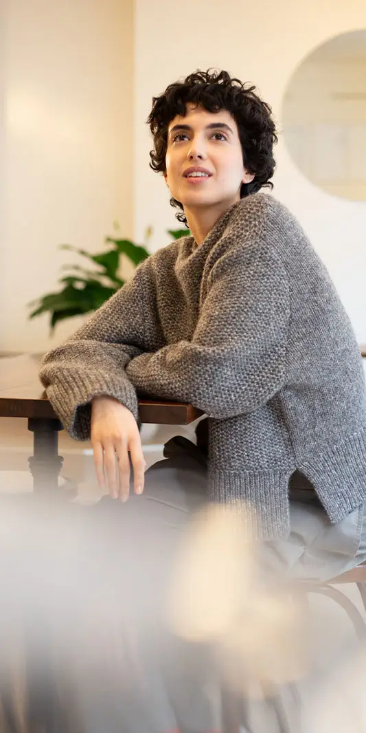 Une femme aux cheveux courts et bouclés porte un pull en tricot gris oversize alors qu'elle est assise à une table à l'intérieur.
