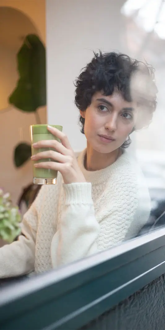 Femme profitant d'un smoothie vert près de la fenêtre.
