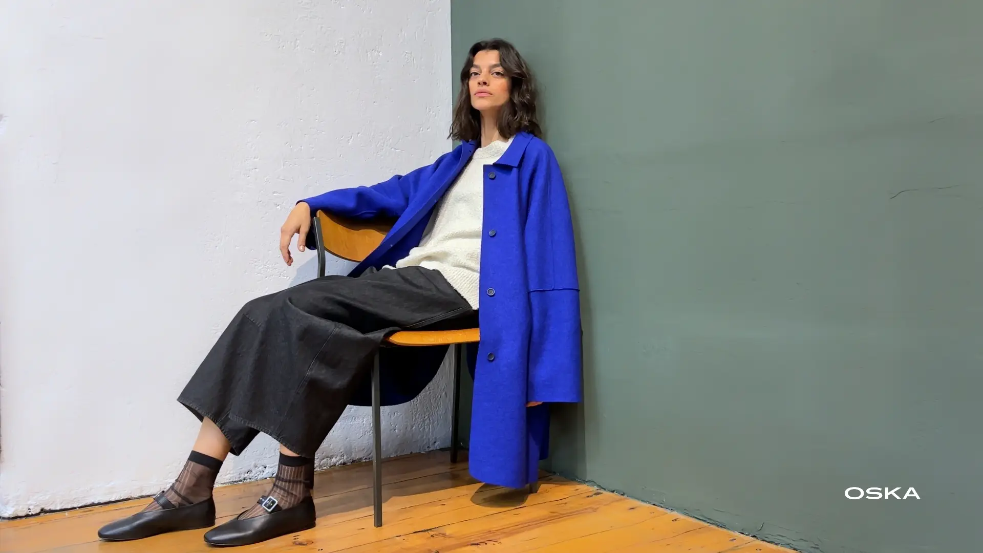Woman in blue coat seated against wall.