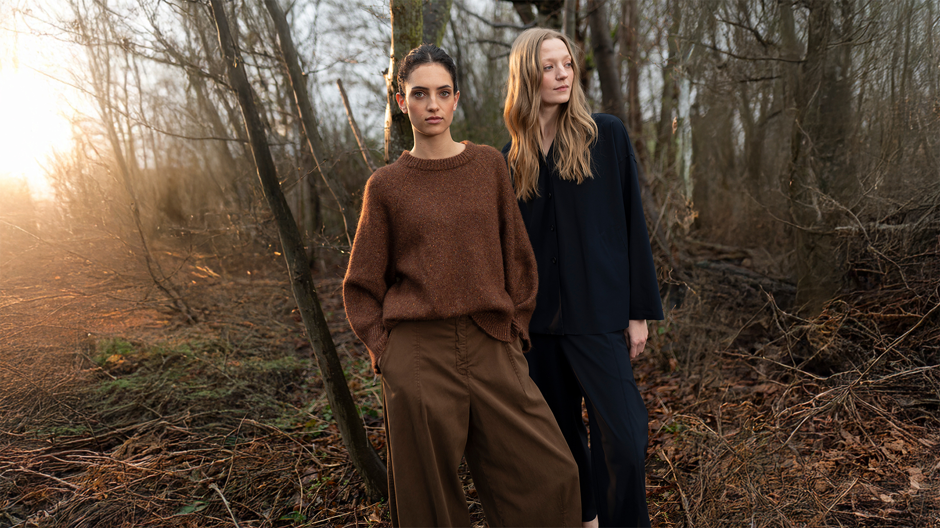 Two women posing in earthy-toned fashion in a sunlit forest.