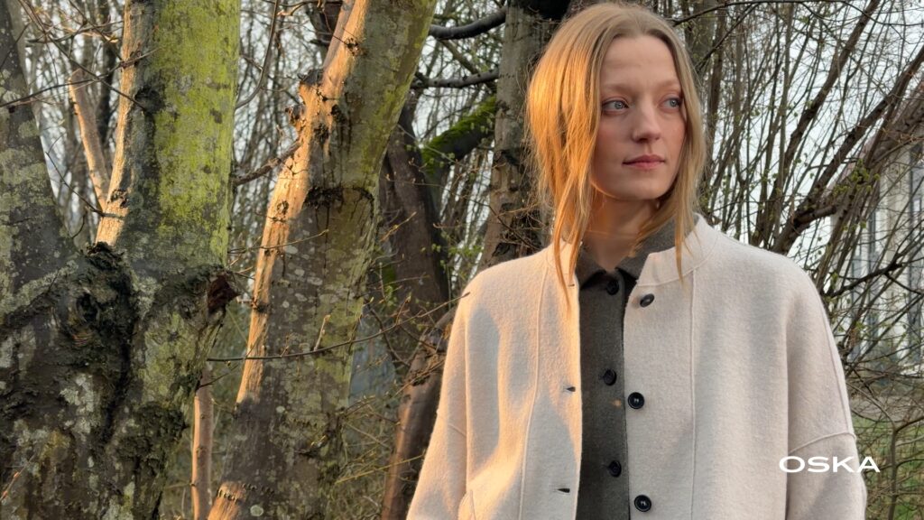 Woman in cream jacket standing among trees at sunset.