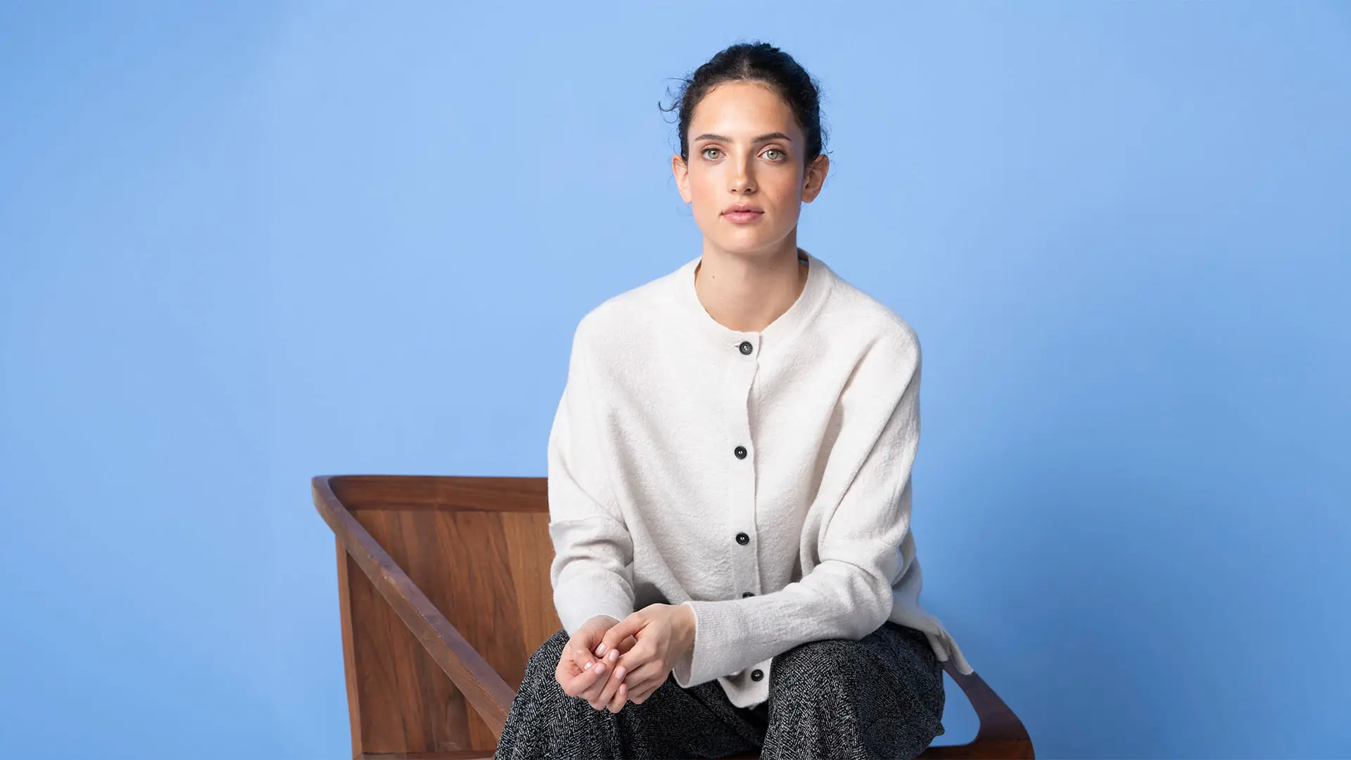 Woman in a cream knitted cardigan sitting on a wooden chair against a blue background